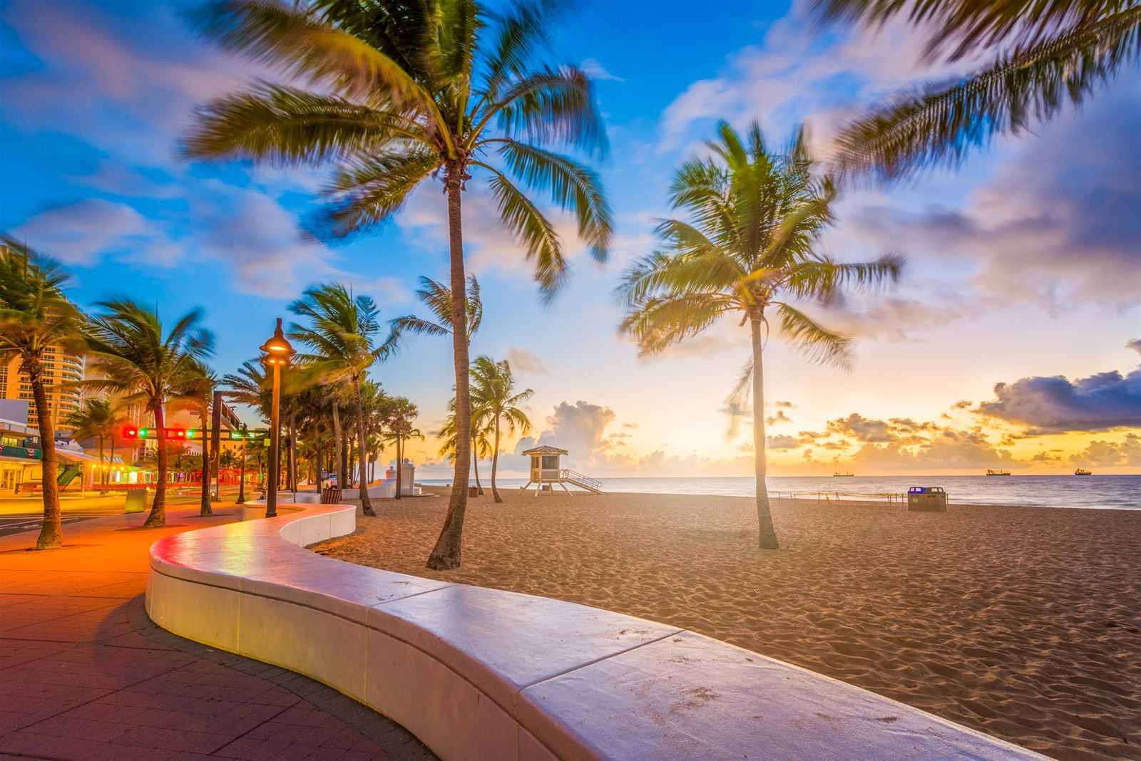 Palm trees blowing over a curving white wall, a beachfront promenade and the sand at sunrise, with a lifeguard stand and ships offshore