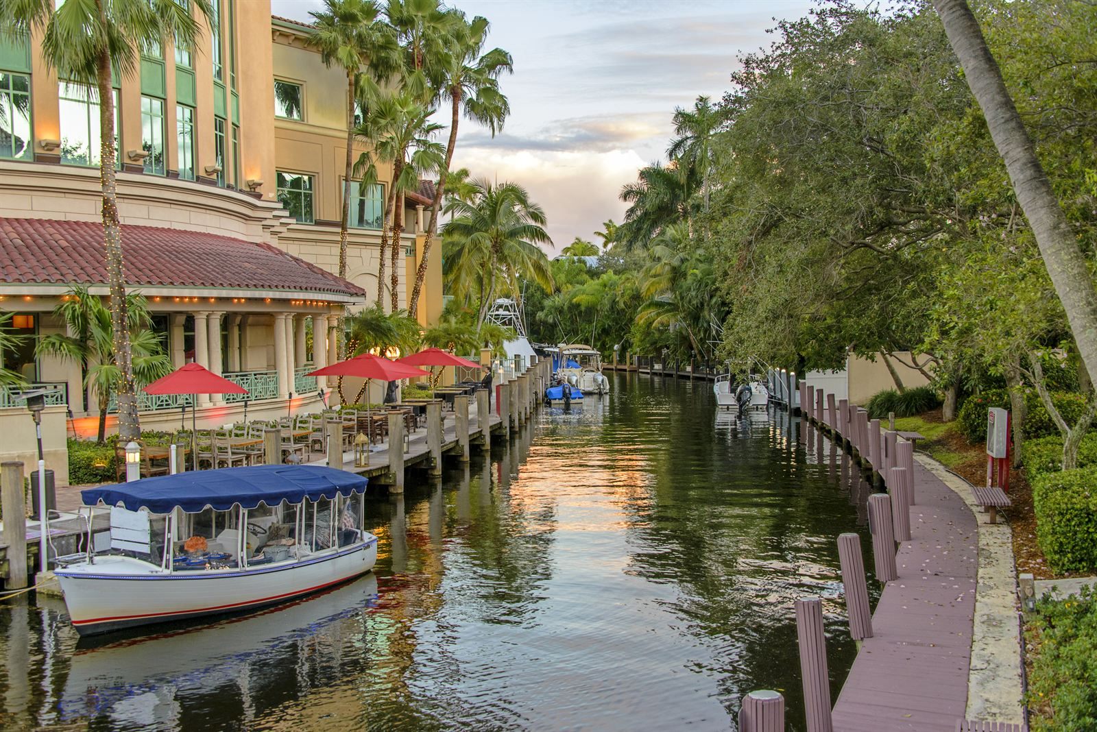 A small covered electric boat docked beside a waterside restaurant patio with red umbrellas on a palm-lined canal at dusk