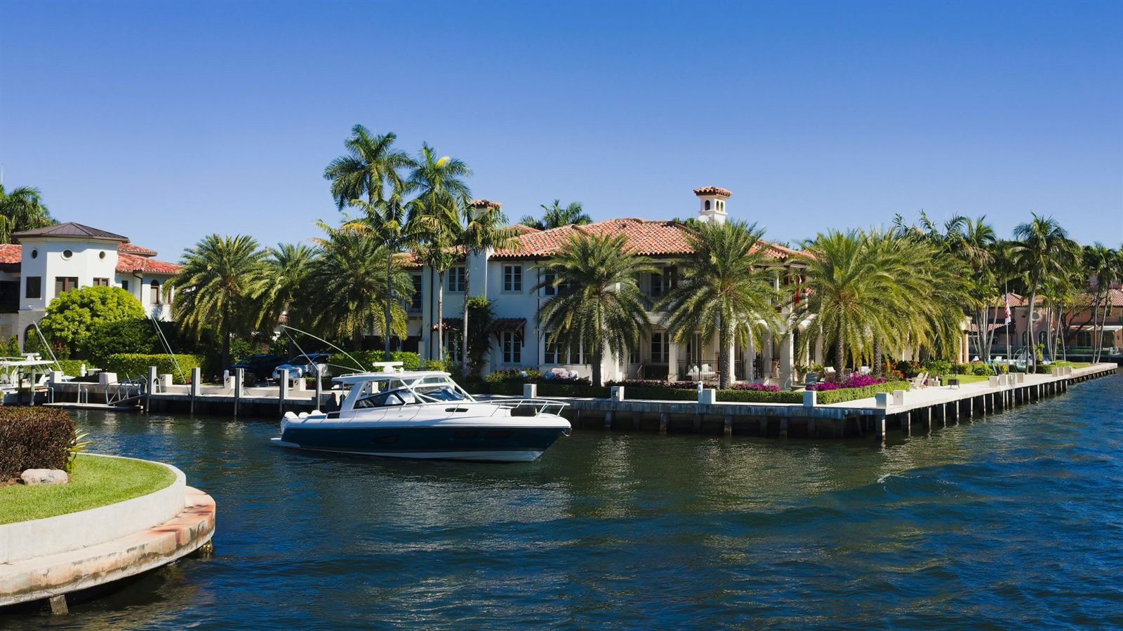 A white motor yacht cruising past a Mediterranean-style waterfront house with a tiled roof and tall palms