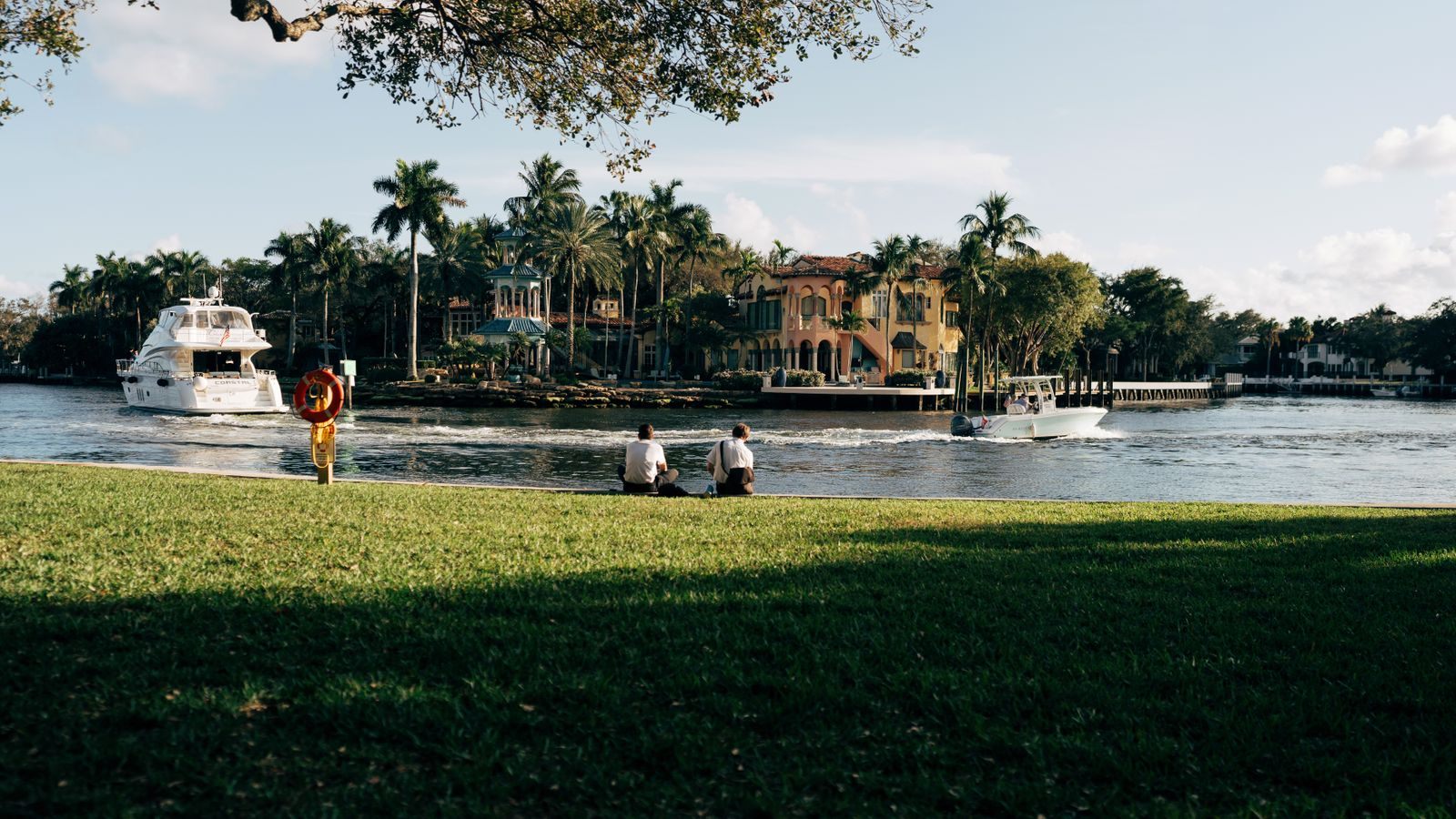 Two people sitting on a riverside lawn as a yacht and a small boat pass a Mediterranean-style house across the water