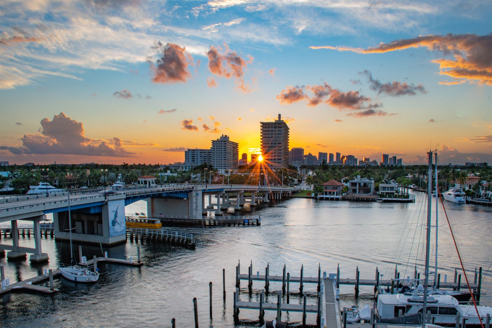 The sun setting behind high-rise towers beyond a bridge, a yellow water taxi and a marina on a Fort Lauderdale waterway