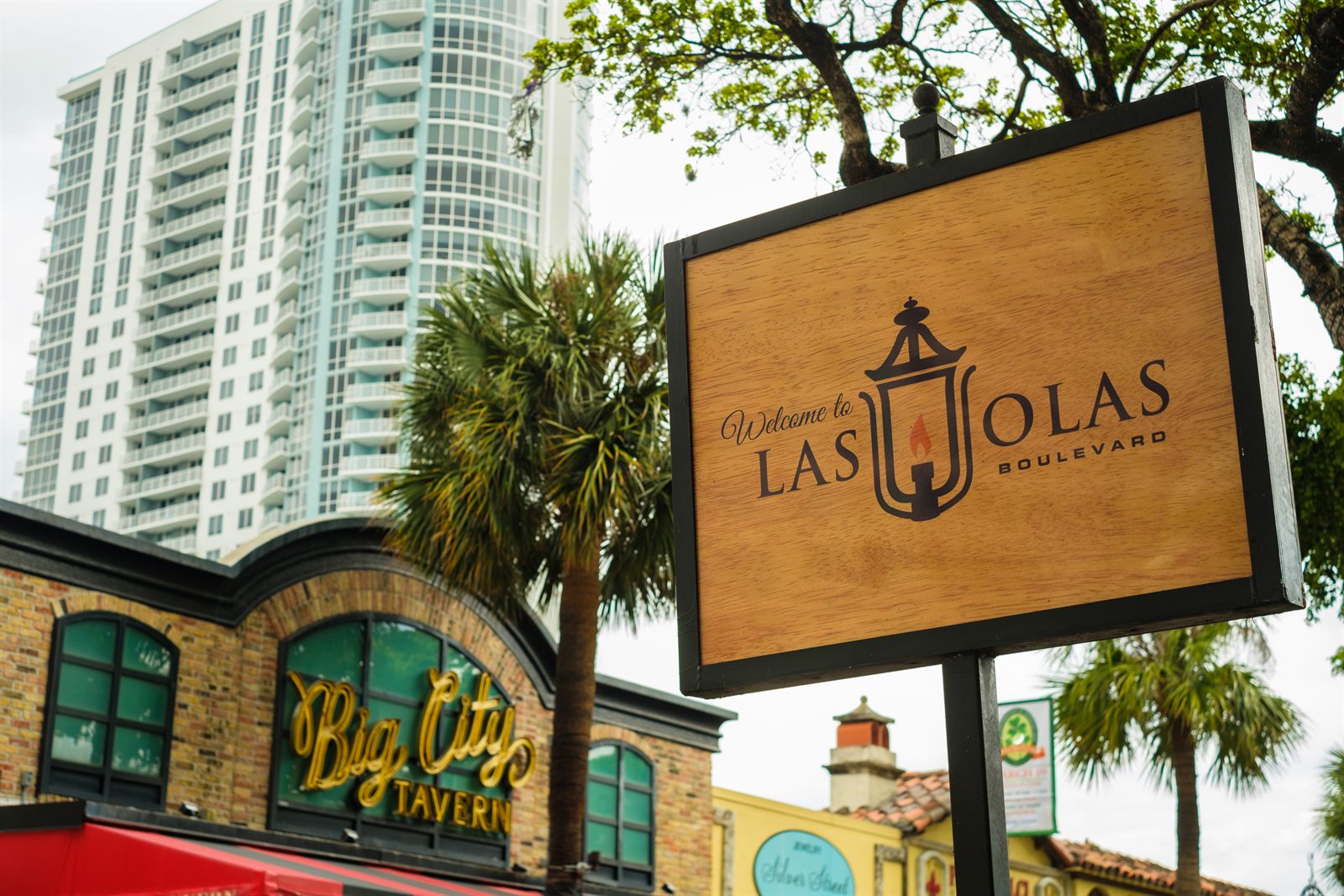A wooden Welcome to Las Olas Boulevard sign beside a brick tavern front, palm trees and a white high-rise tower