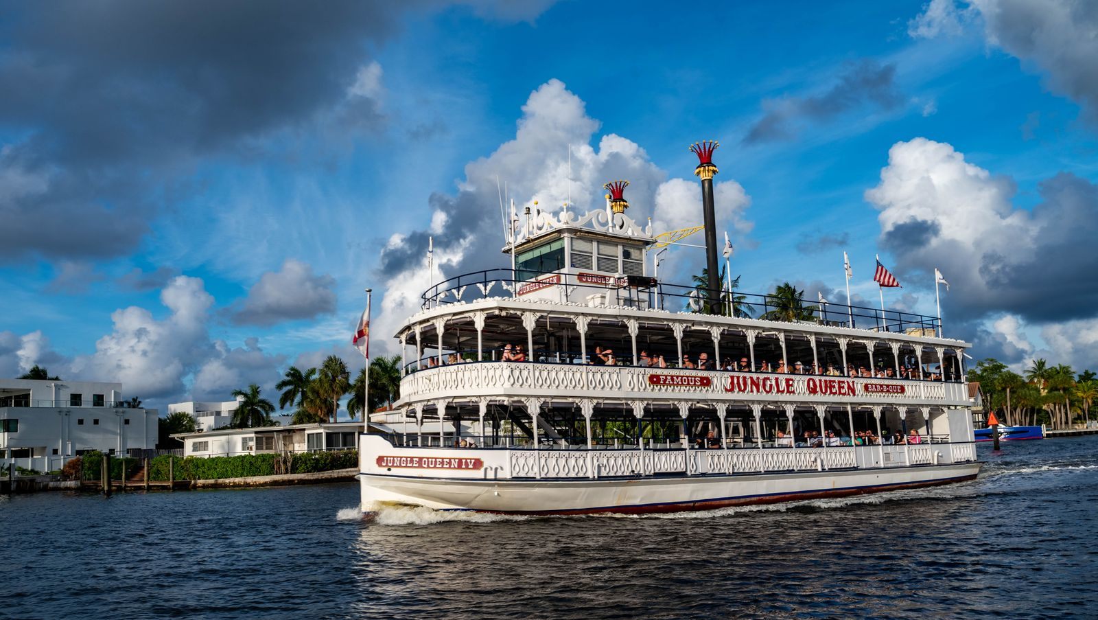 The white Jungle Queen IV riverboat carrying passengers on its upper and lower decks under a blue sky with tall clouds